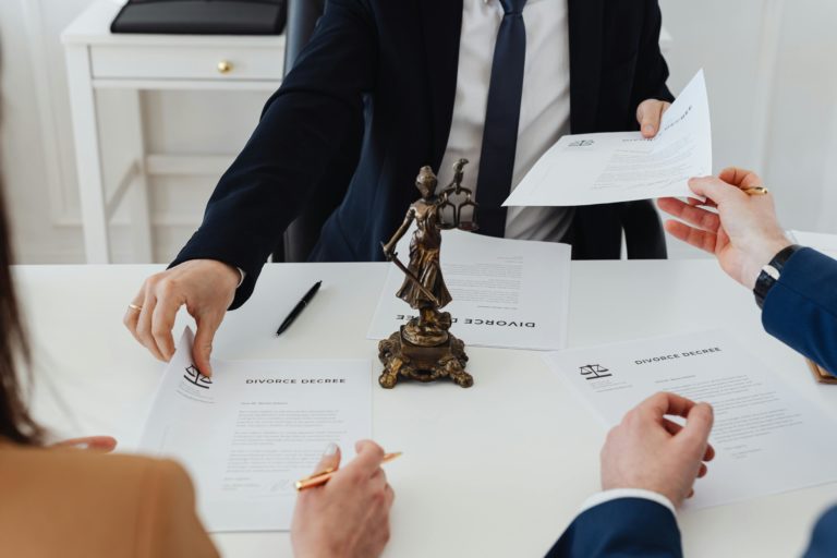 Three people in formal attire handling documents at a table with a Lady Justice statue.